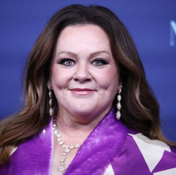melissa mccarthy smiles at the camera, her hair is down, she wears a purple and white outfit and a white necklace and earrings