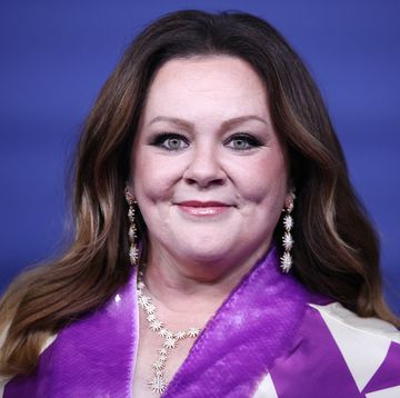 melissa mccarthy smiles at the camera, her hair is down, she wears a purple and white outfit and a white necklace and earrings