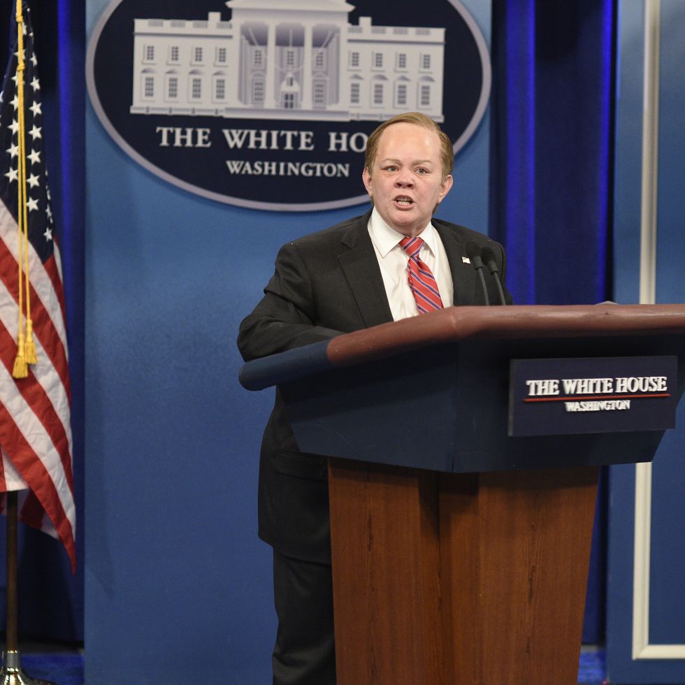 melissa mccarthy in character as sean spicer, she is standing in a fake white house briefing room at the podium and speaking, she wears a dark suit with white collared shirt and red tie with blue and white stripes