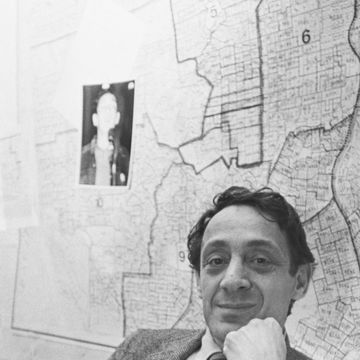 a man in a suit jacket striped tie and collared shirt smiles at the camera as he leans his head against his fist, a map covers the wall behind him