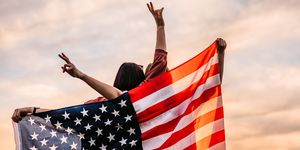two, young, females, covering themselves with usa flag standing on the meadow at sunset rear view