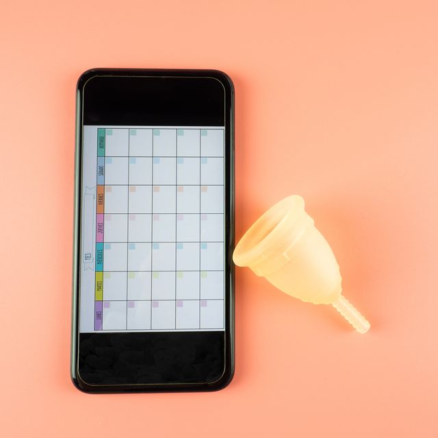a menstrual cup next to a smartphone with a menstrual calendar mobile app on a pink background