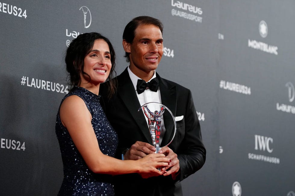 xisca perello and rafael nadal smiling as they simultaneously hold a trophy, she wears a sparkling sleeveless dress, he wears a black suit jacket and bow tie with a white collared shirt