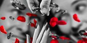 a black and white image of engagement rings floating towards a hand with a couple in the background and red rose petals