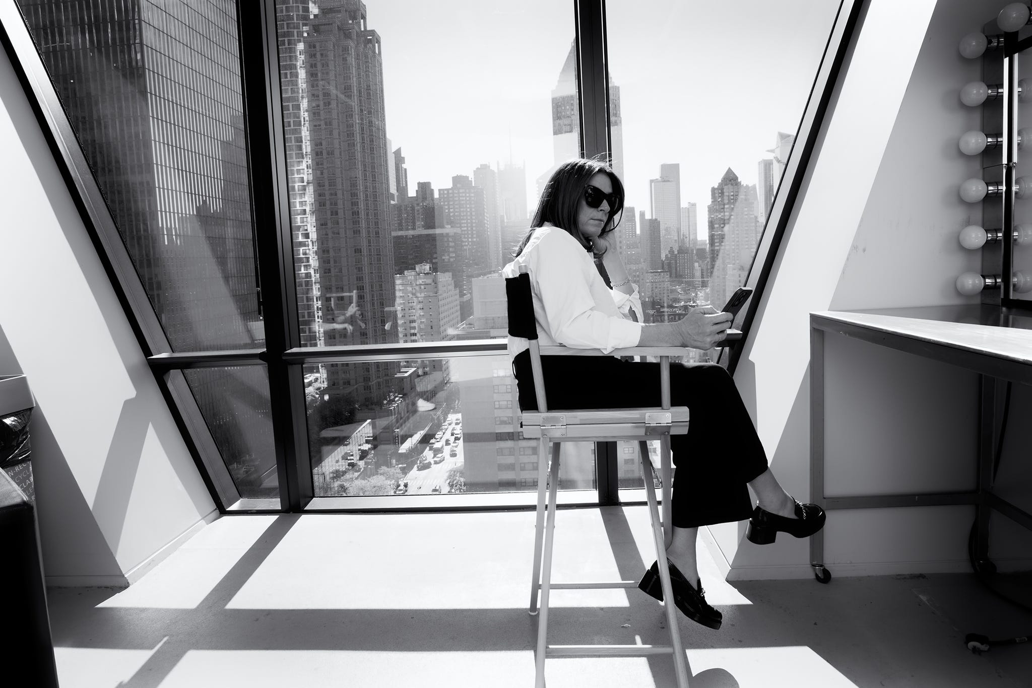 a person sits in a directors chair holding a smartphone and appearing to be engaged in a phone conversation they are dressed in a white shirt and black pants the setting is an office or studio with large windows showcasing a panoramic view of a city skyline characterized by tall buildings a vanity mirror with lights is visible in the background, adding a professional or studio like ambience