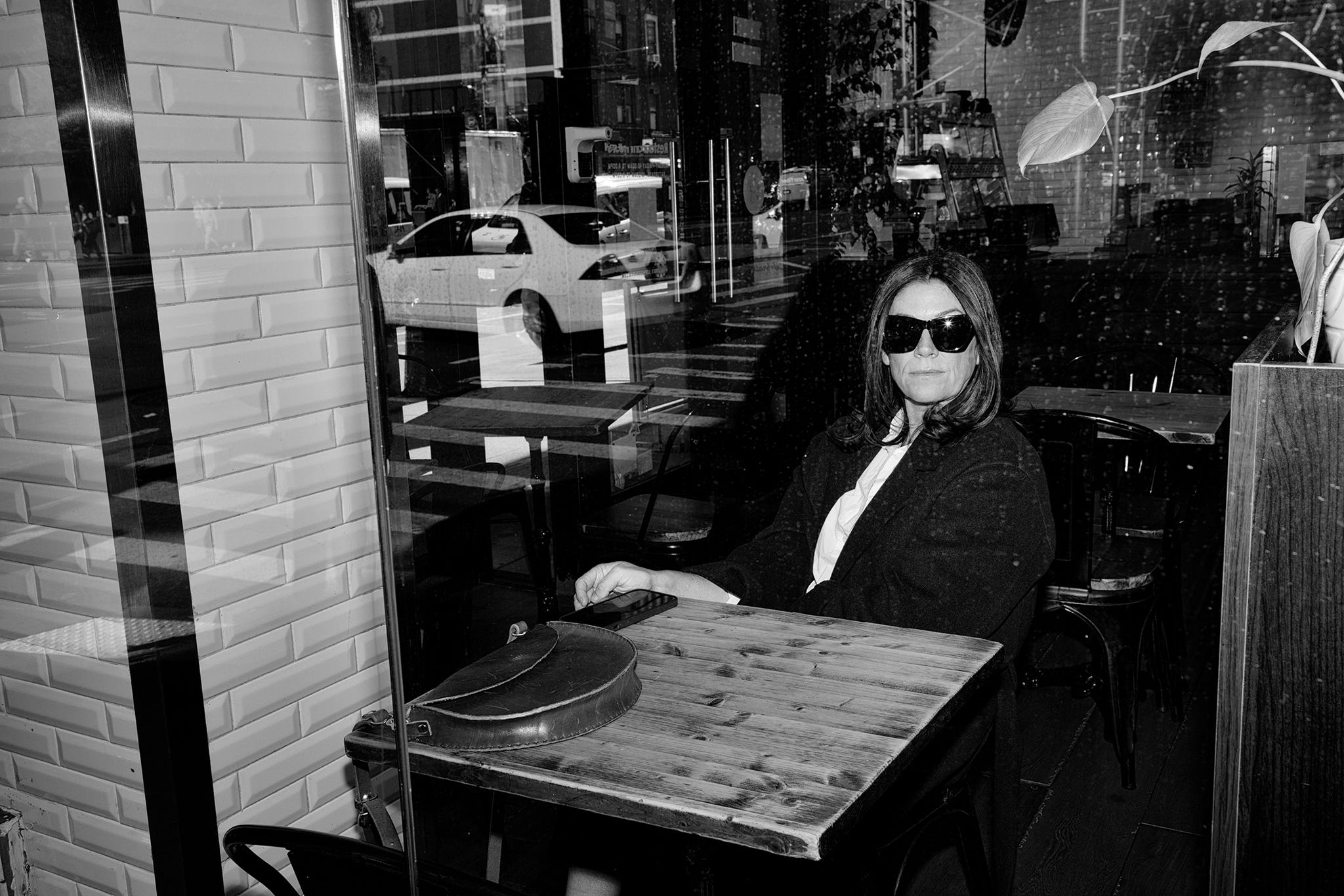 an indoor cafe a cute scene featuring a person seated at a wooden table the table has a rounded bag positioned on it and the individual is dressed in a dark garment with a white shirt underneath the background displays glass panes reflecting the exterior street and other cafe a cute elements contributing to a cozy urban atmosphere