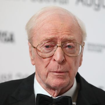 michael caine stands in front of a white blurred background and wears a black suit jacket, black bowtie, white collared shirt and large wire rimmed glasses, he looks to the left of the camera with a solemn expression on his face