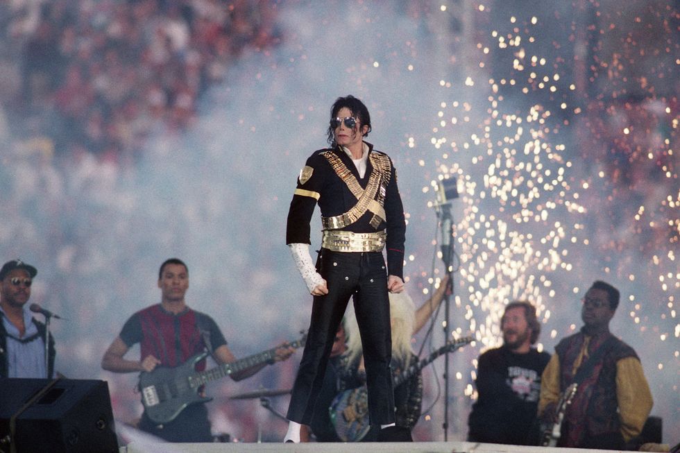 michael jackson stands on stage at the super bowl xxxviii halftime show, he wears a black outfit with gold accents and a white sleeve up to his right elbow, behind him are musicians, pyrotechnics and the crowd