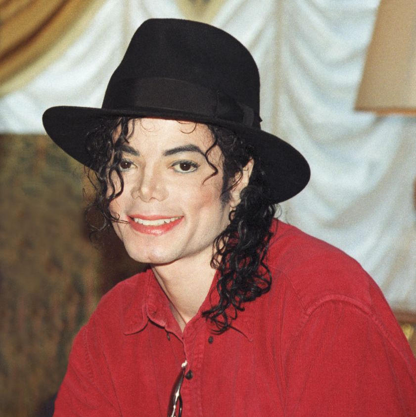 michael jackson smiles in a 1996 photo, he is wearing a red button up shirt with sunglasses tucked into his shirt and a black wide brimmed hat, his black curly hair is shoulder length and frames his face