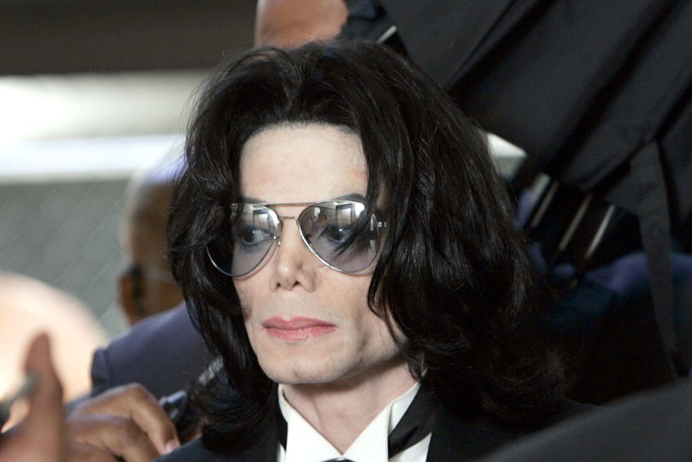 michael jackson wears blue tinted sunglasses, a black suit and tie, and a neutral expression, he is standing in a crowd