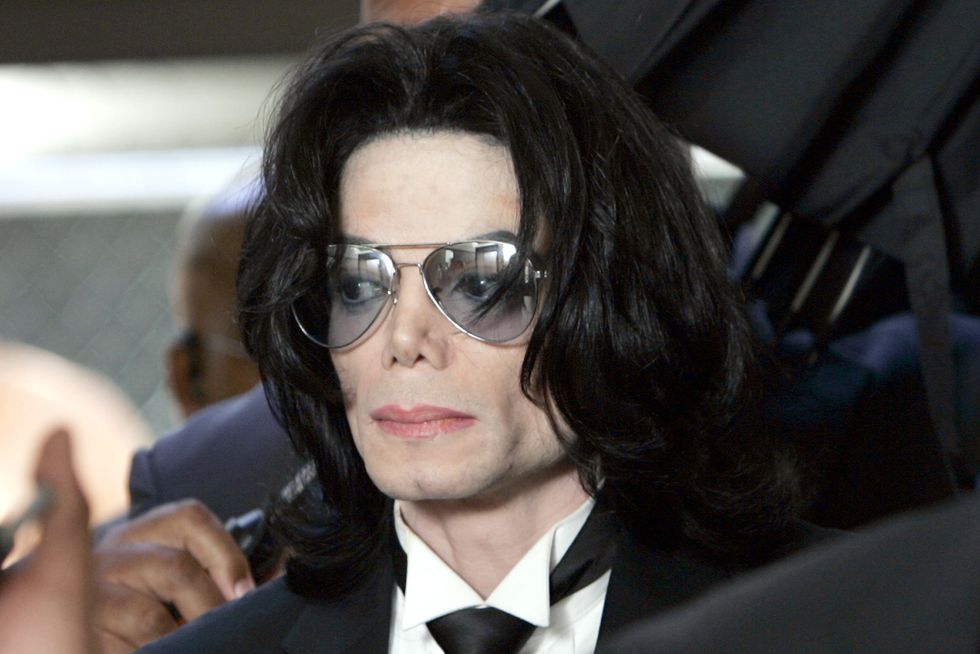 michael jackson wears blue tinted sunglasses, a black suit and tie, and a neutral expression, he is standing in a crowd