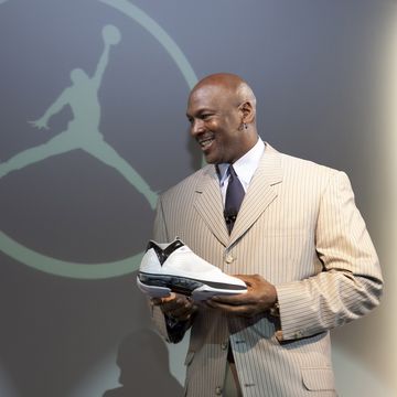 michael jordan, wearing a tan pinstriped suit and black tie, smiles and holds a sneaker while standing in front of the nike air jordan logo