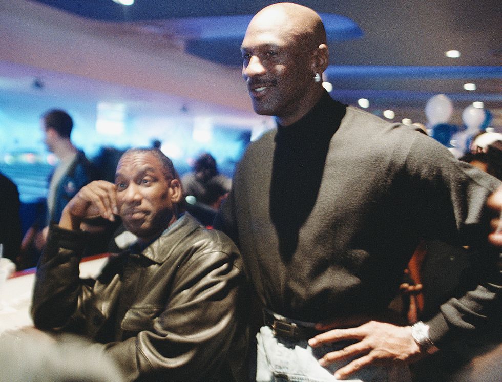 howard white, wearing a black leather jacket, sits next to a standing michael jordan, wearing a black suit, in the middle of a crowded room