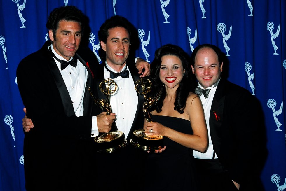 michael richards, jerry seinfeld, julia louis dreyfus, and jason alexander pose for a photo in front of an blue emmy award backdrop, richards and louis dreyfus are holding emmy statues as all four hug and smile