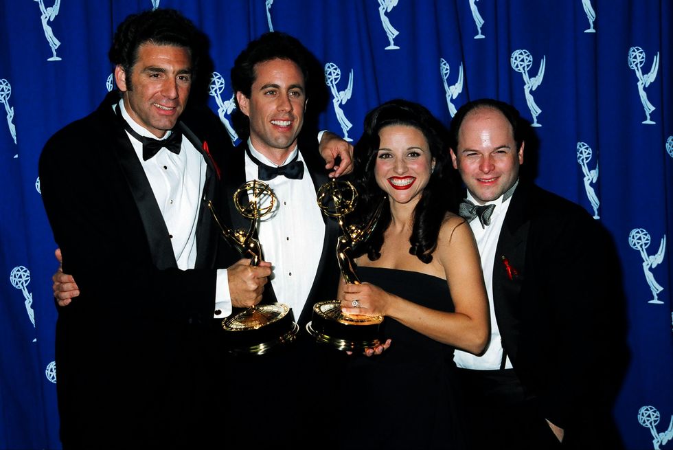 michael richards, jerry seinfeld, julia louis dreyfus, and jason alexander pose for a photo in front of an blue emmy award backdrop, richards and louis dreyfus are holding emmy statues as all four hug and smile