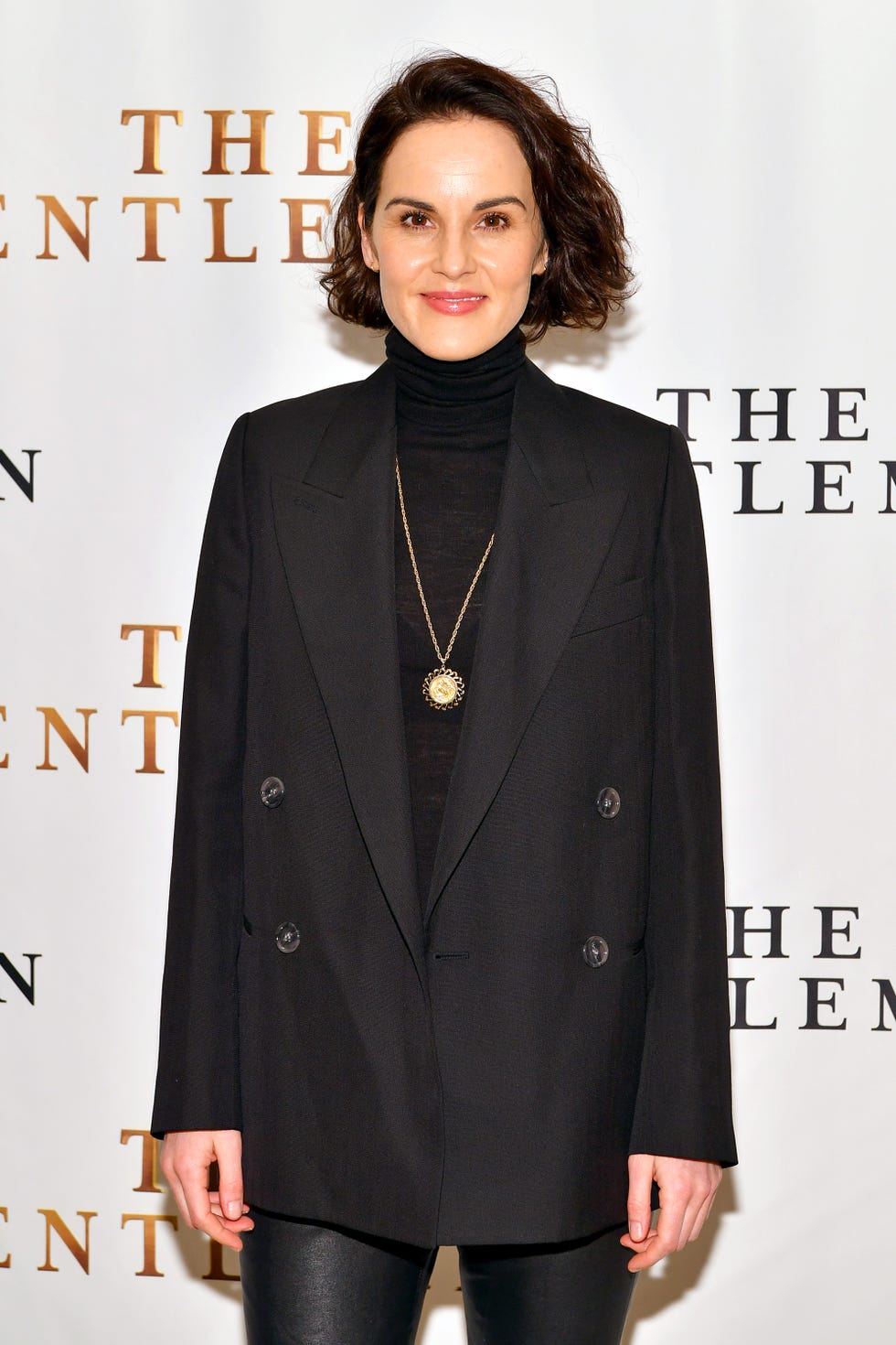 michelle dockery is a young woman wearing a black turtle neck sweater, black suit jacket and black leggings with a gold chain, she is smiling at the camera with her short brown hair in a wavy bob