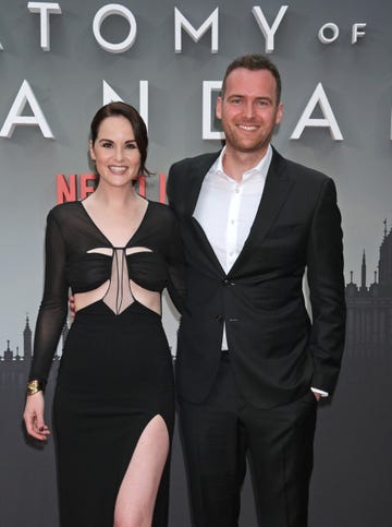 michelle dockery and jasper waller bridge stand with arms around each other and smiling at the camera, she wears a long back dress and he a black suit