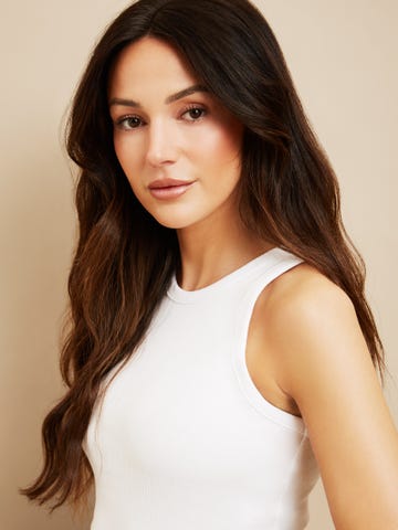 woman with long wavy hair wearing a sleeveless white top against a neutral background