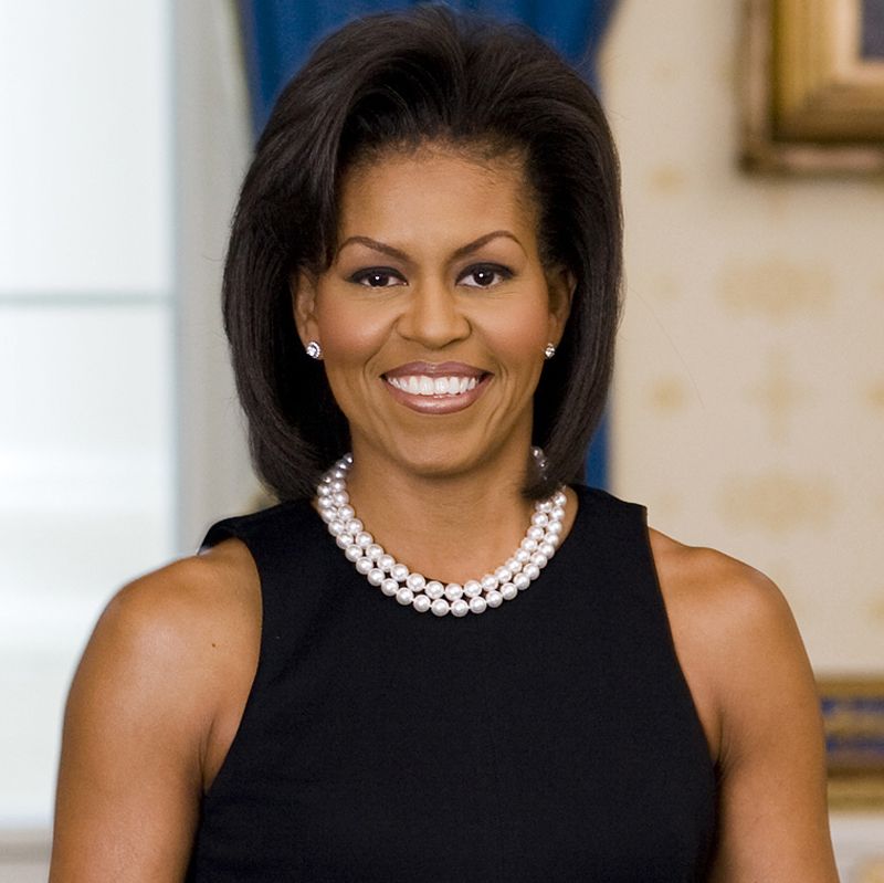 first lady michelle obama in a black dress and pearl necklace, smiling at the camera
