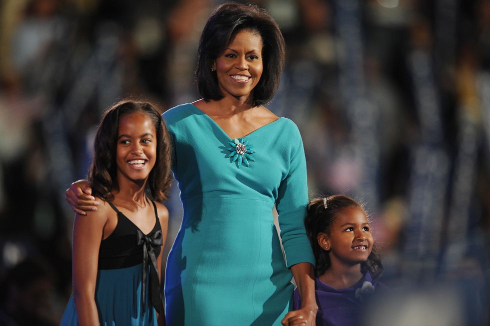 malia obama, michelle obama, and sasha obama smile as they look various directions, malia is wearing a black and blue dress with a bow in the front, michelle is wearing a teal dress with three quarter length sleeves and a flower broach, sasha is wearing a purple top