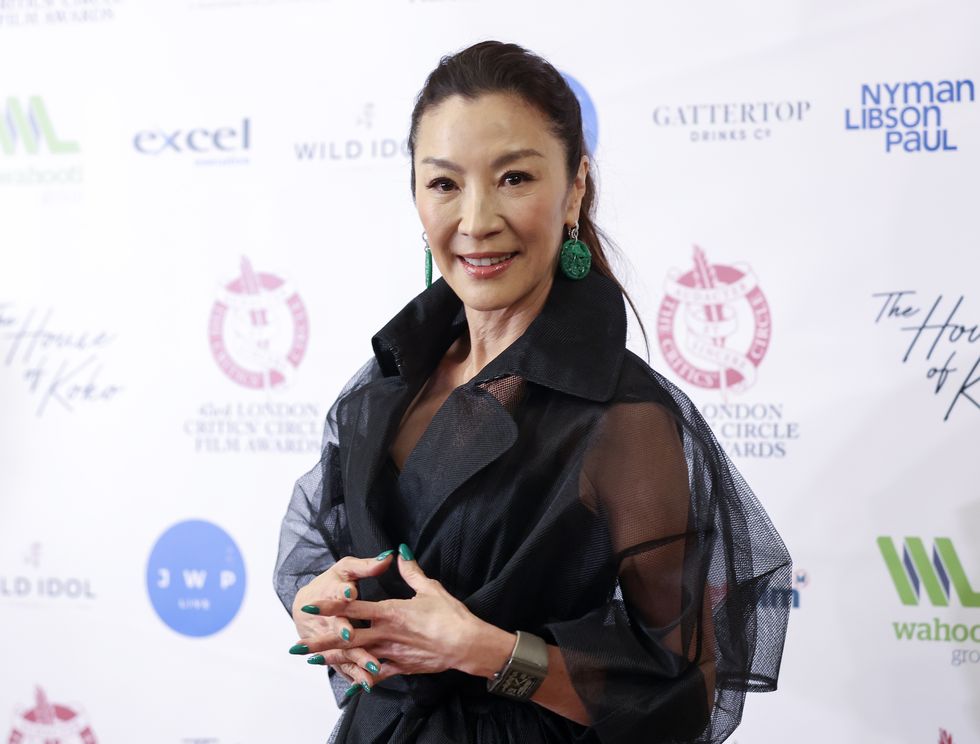 michelle yeoh, wearing a black dress, folds her hands and smiles for a publicity photo