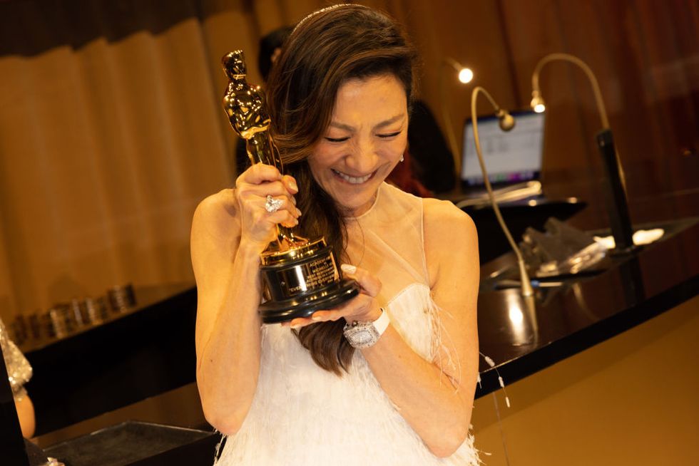 michelle yeoh, smiling and looking down with her eyes closed, holding an academy award, wearing a white dress, and standing in front of a table in a darkened room