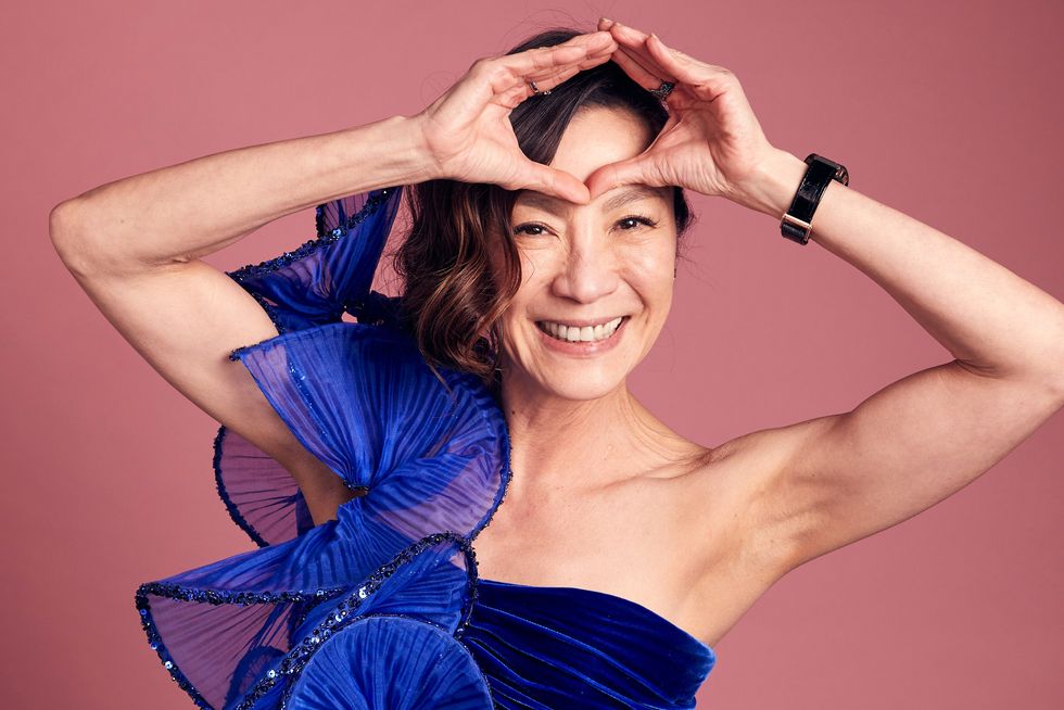michelle yeoh wearing a blue dress, smiling and holding her hands on her forehead in a circle shape