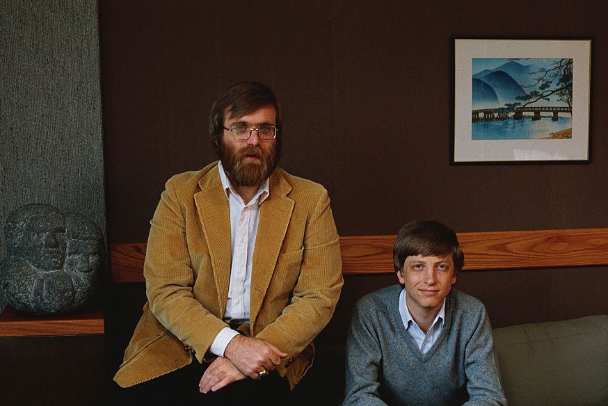paul allen and bill gates sit on a couch and pose for a portrait, allen wears a corduroy blazer, white collared shirt and dark pants, gates wears a gray sweater, blue collared shirt and dark pants