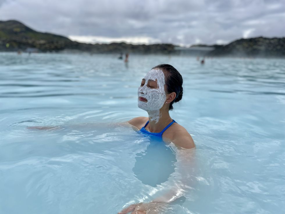 mid adult asian woman relaxing in blue lagoon on iceland, female with face smeared with white healing mud