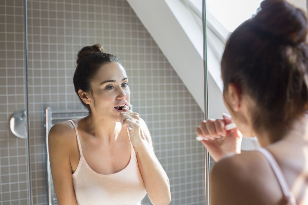 Mid adult woman brushing teeth in bathroom mirror.
