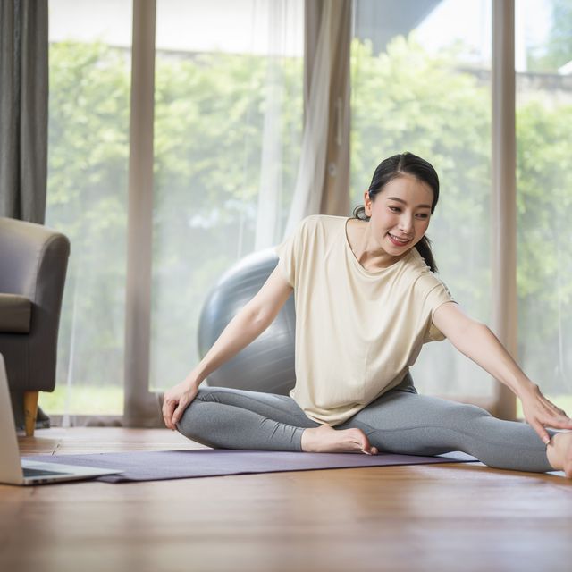 mid adult woman sitting on exercise mat and stretching exercise while learning live streaming tutorial at home healthy lifestyle and work life balance concepts
