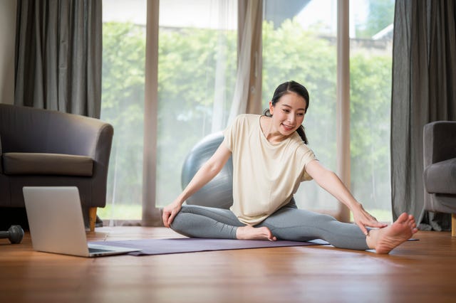 mid adult woman sitting on exercise mat and stretching exercise while learning live streaming tutorial at home healthy lifestyle and work life balance concepts