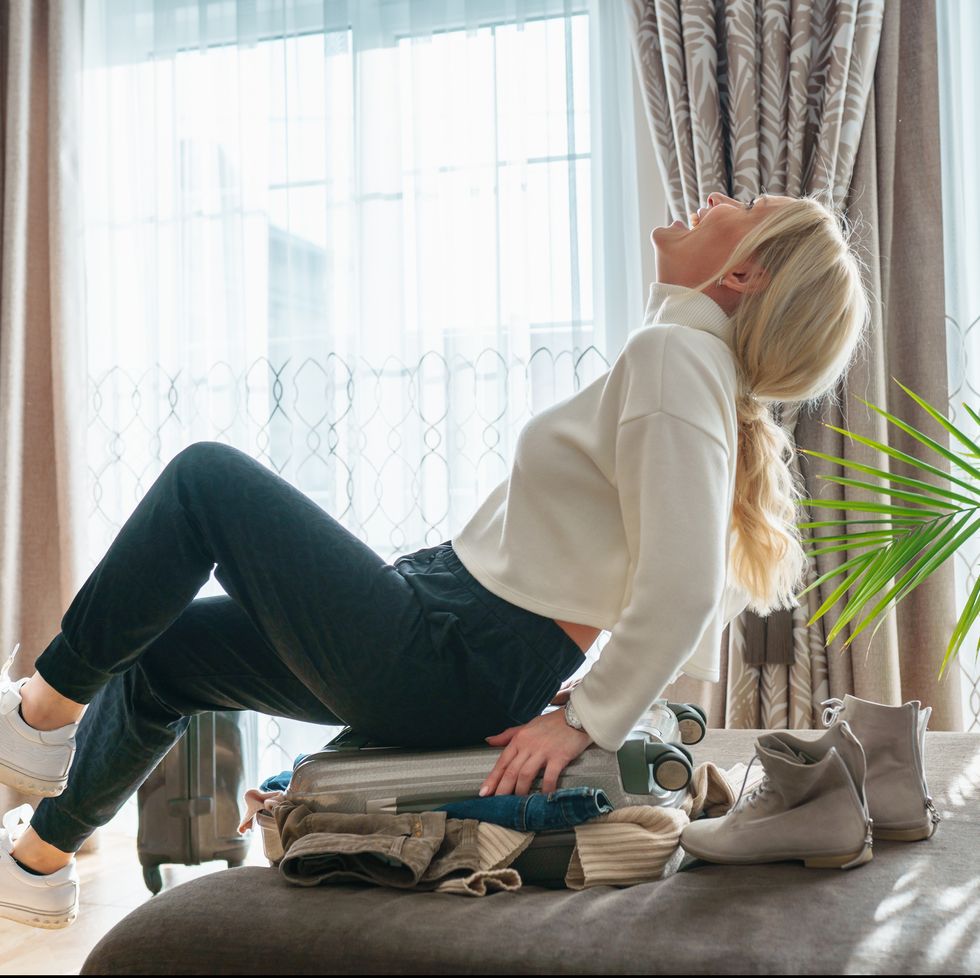middle age blonde smiling woman in white sweater trying to close suitcase full of clothes in living room at home female sit on a suitcase and laughing