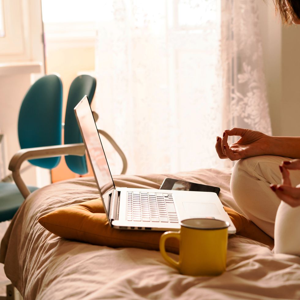 a middle aged woman in white jeans and a yellow sweater sitting on the bed in a yoga pose in front of a laptop and a cup of coffee