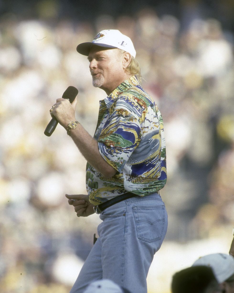 a man in a hawaiian shirt and jeans sings into a microphone he holds in one hand