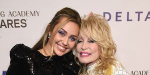 miley cyrus and dolly parton hug and smile for a photo together, cyrus wears a sequined black long sleeve dress, parton wears a white long sleeve dress with a gold studded pattern