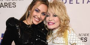 miley cyrus and dolly parton hug and smile for a photo together, cyrus wears a sequined black long sleeve dress, parton wears a white long sleeve dress with a gold studded pattern