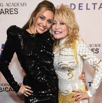 miley cyrus and dolly parton hug and smile for a photo together, cyrus wears a sequined black long sleeve dress, parton wears a white long sleeve dress with a gold studded pattern