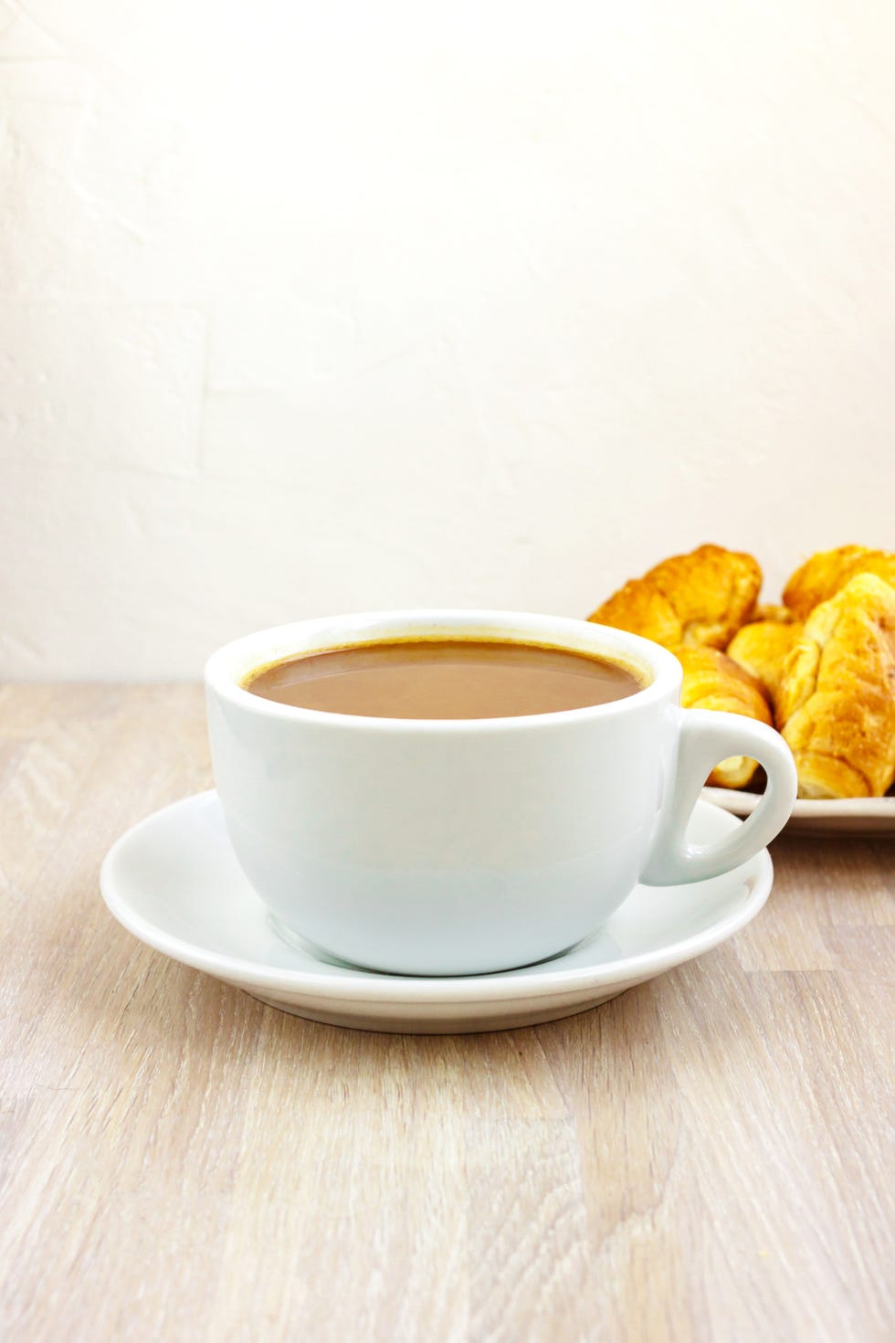 milk coffee cup with saucer and a croissants on light wooden background close up breakfast concept