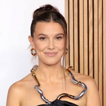 millie bobby brown, a young woman, wears her hair up and a black strapless gown with a sculpted metal neck piece and large hoop earings while posing for photographers at the glamour women of the year 2023 arrivals