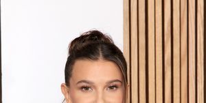 millie bobby brown, a young woman, wears her hair up and a black strapless gown with a sculpted metal neck piece and large hoop earings while posing for photographers at the glamour women of the year 2023 arrivals