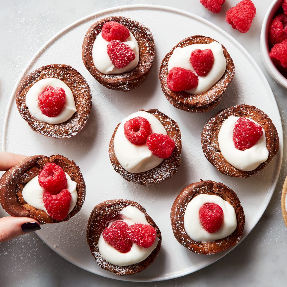 miniature chocolate flavored dutch baby pancakes sitting in the wells of a muffin tin and garnished with whipped cream, raspberries, and powdered sugar
