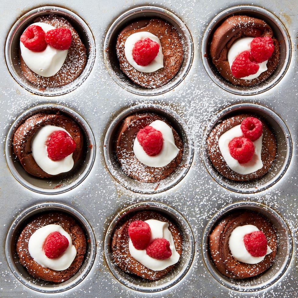 miniature chocolate flavored dutch baby pancakes sitting in the wells of a muffin tin and garnished with whipped cream, raspberries, and powdered sugar