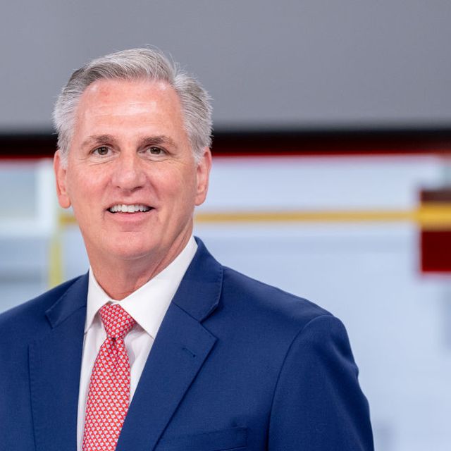 kevin mccarthy smiles at the camera, he wears a blue suit jacket, white collared shirt, and red tie with white polka dots