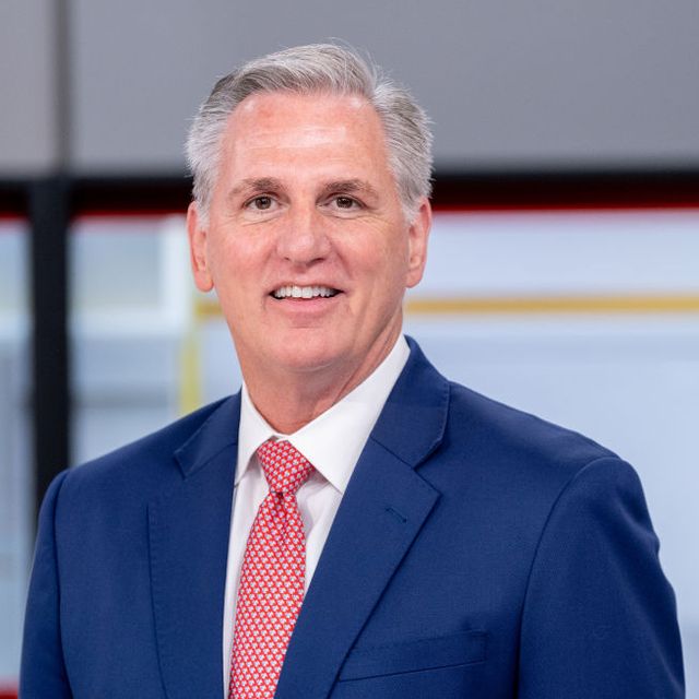kevin mccarthy smiles at the camera, he wears a blue suit jacket, white collared shirt, and red tie with white polka dots
