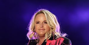 miranda lambert smiles while performing, she wears a black western outfit with pink fringe and embellishments