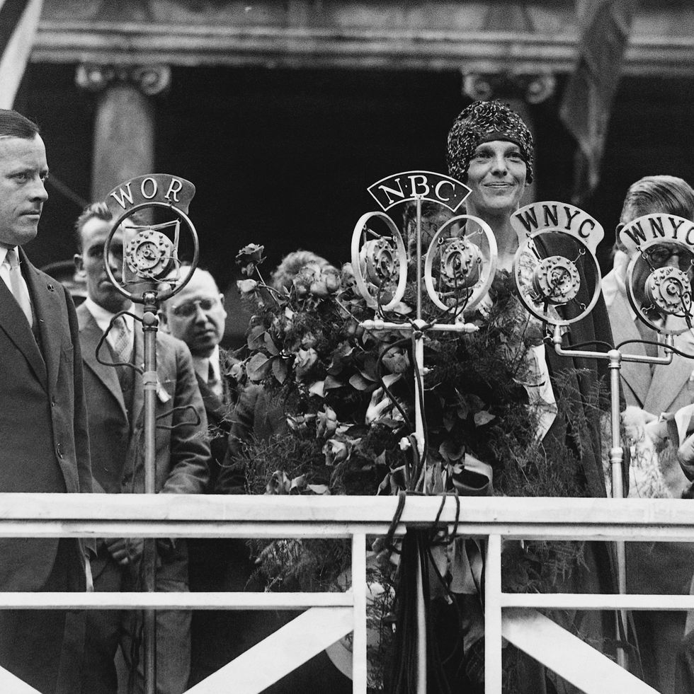 amelia earhart smiles and stands behind several microphones, she holds a large bouquet and wears a cap and dress