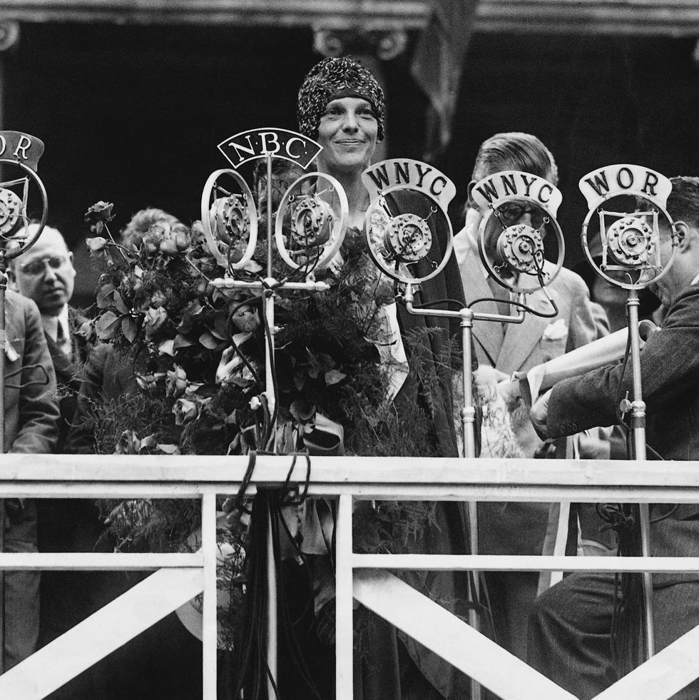 amelia earhart smiles and stands behind several microphones, she holds a large bouquet and wears a cap and dress