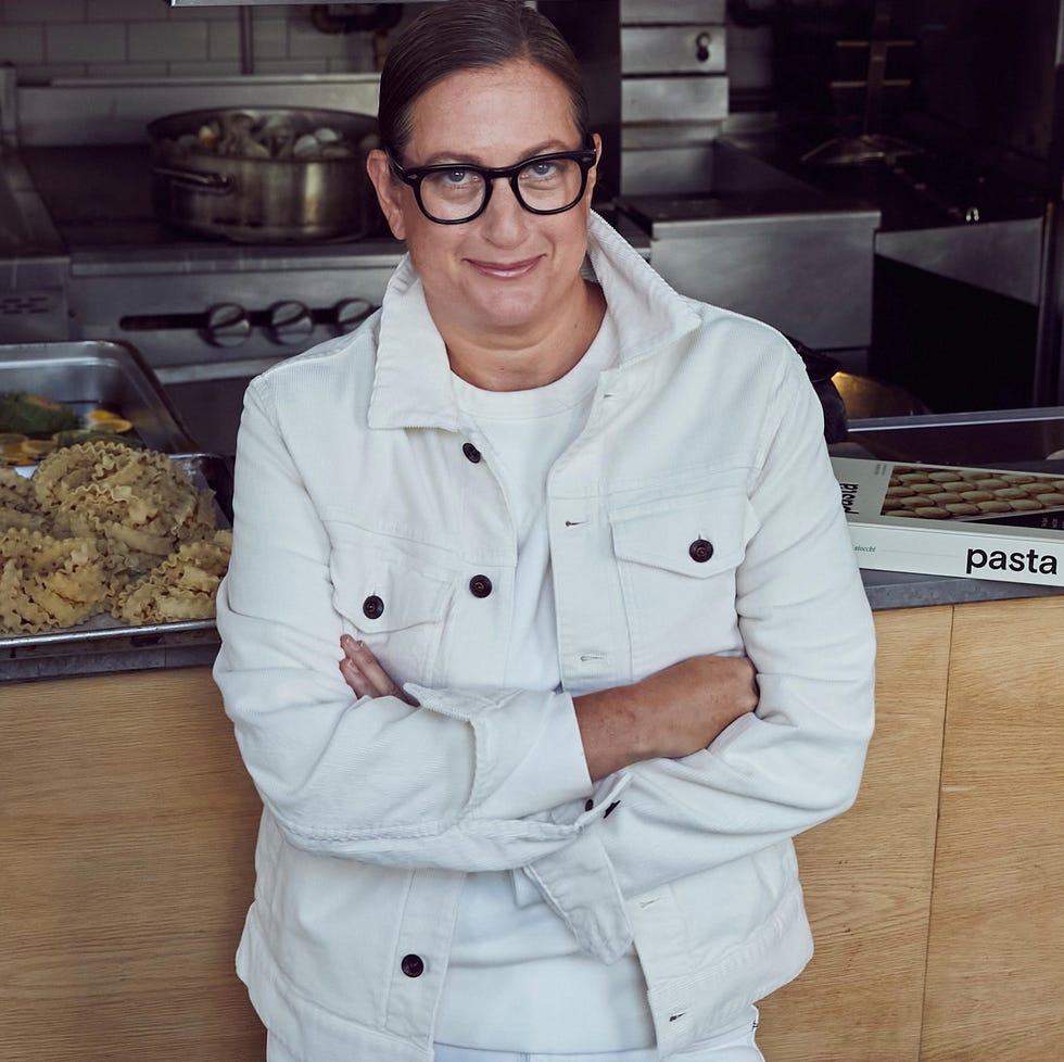 a person in a white jacket stands in front of a kitchen with pasta in the background