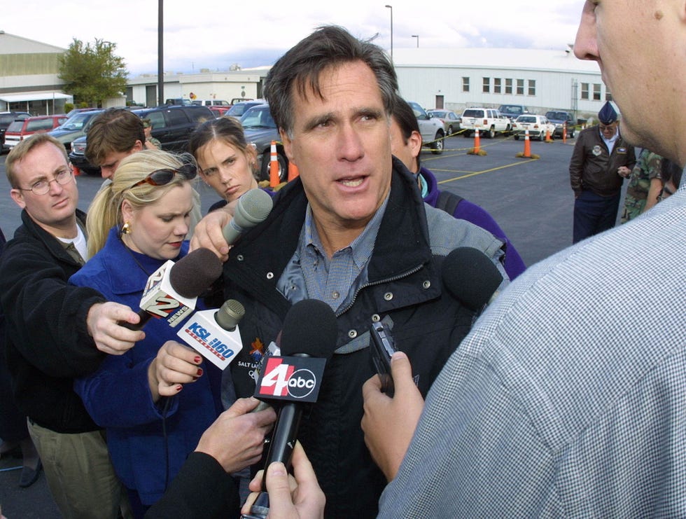 mitt romney wearing a jacket, talking to reporters holding several microphones in front of him as they stand in a parking lot outside